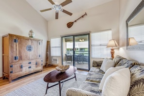 A living room with a wooden cabinet, a ceiling fan, a dining area with a table and chairs, and a large window with blinds. - Relax in Kihei | Ocean View Condo, Pool, Hot Tub, Walk to Beach (Kihei)
