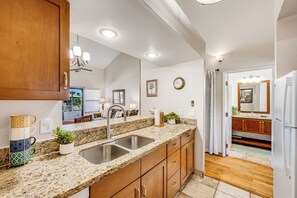 A kitchen with granite countertops, stainless steel sink, wooden cabinets, and a view into the dining area. - Relax in Kihei | Ocean View Condo, Pool, Hot Tub, Walk to Beach (Kihei)