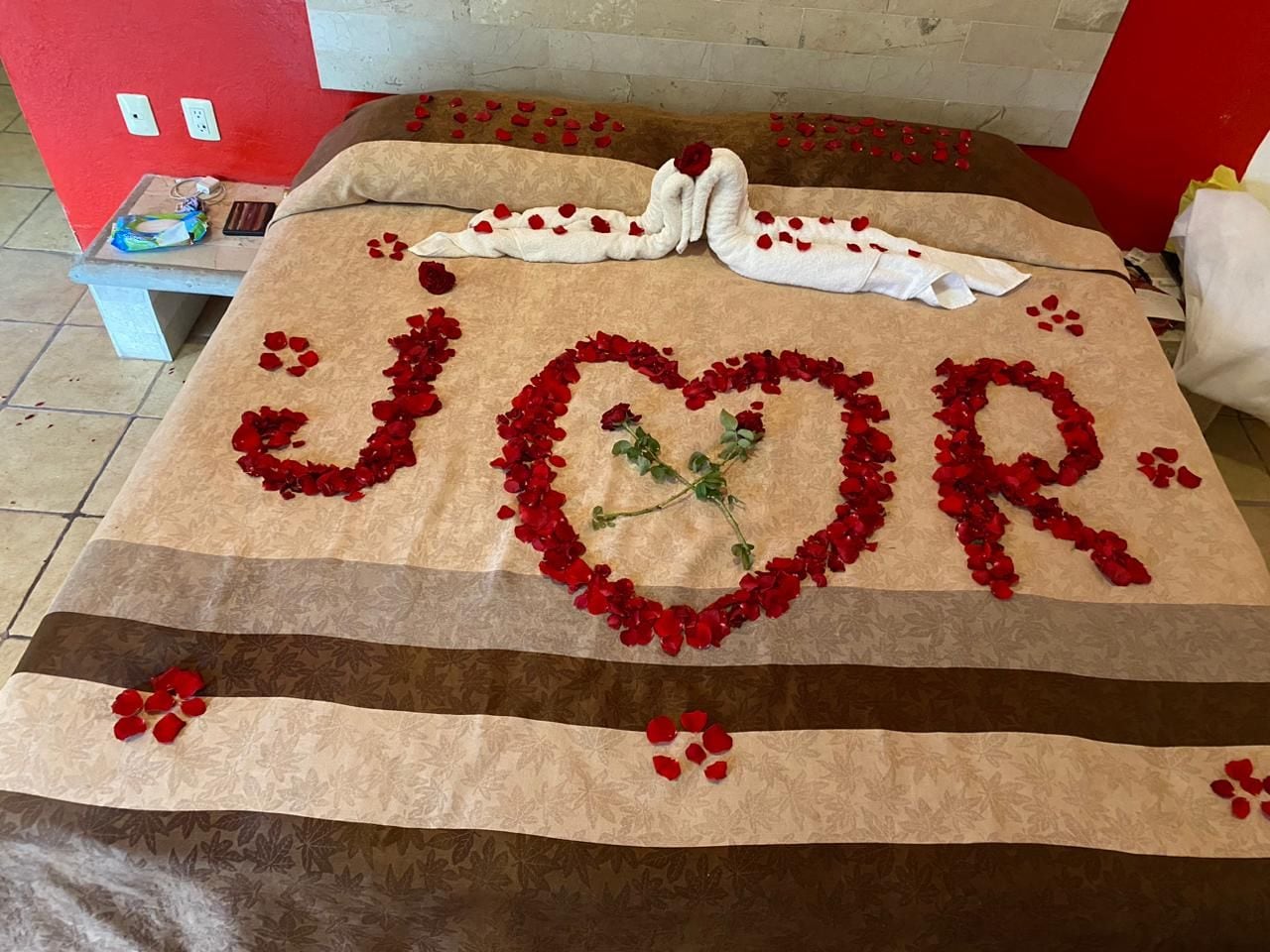 Una cama con un arreglo de pétalos de rosa roja formando la palabra 'JOUR' y una paloma blanca con una rosa roja sobre la almohada.