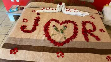 Una cama con un arreglo de pétalos de rosa roja formando la palabra 'JOUR' y una paloma blanca con una rosa roja sobre la almohada.