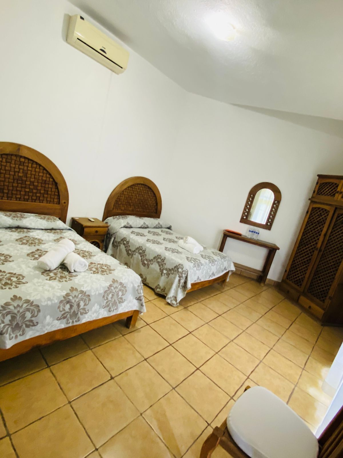 A hotel room with two beds, a bedside table, a mirror, a wardrobe, and an air conditioning unit.