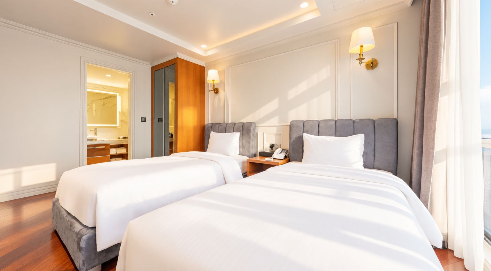 Habitación de hotel con dos camas, una mesita de noche con lámpara, un espejo y vista al mar.