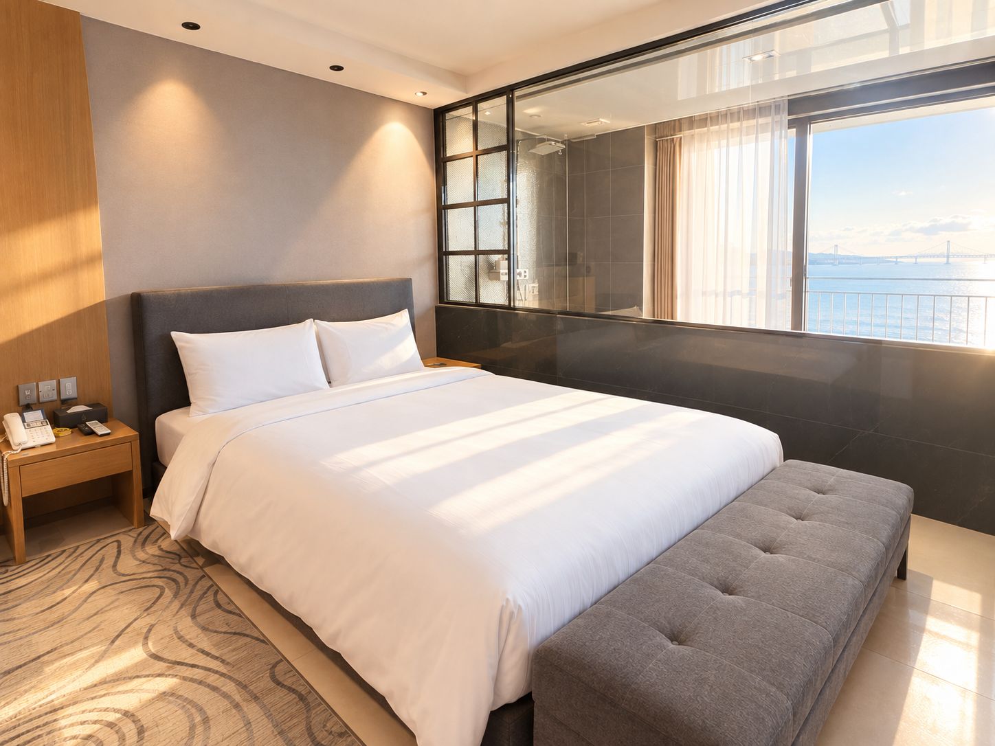 A modern hotel room with a large bed, a bedside table with a phone and a lamp, a grey bench, and a large window with a view of a bridge and water.
