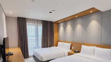 A hotel room with two beds, a desk, a TV, and a large window with curtains.