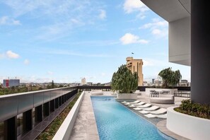 A rooftop swimming pool with a view of the city skyline, outdoor seating, and a tall building in the background. - Queen's Wharf Residences Luxury 2 Beds 1 Car (Brisbane)