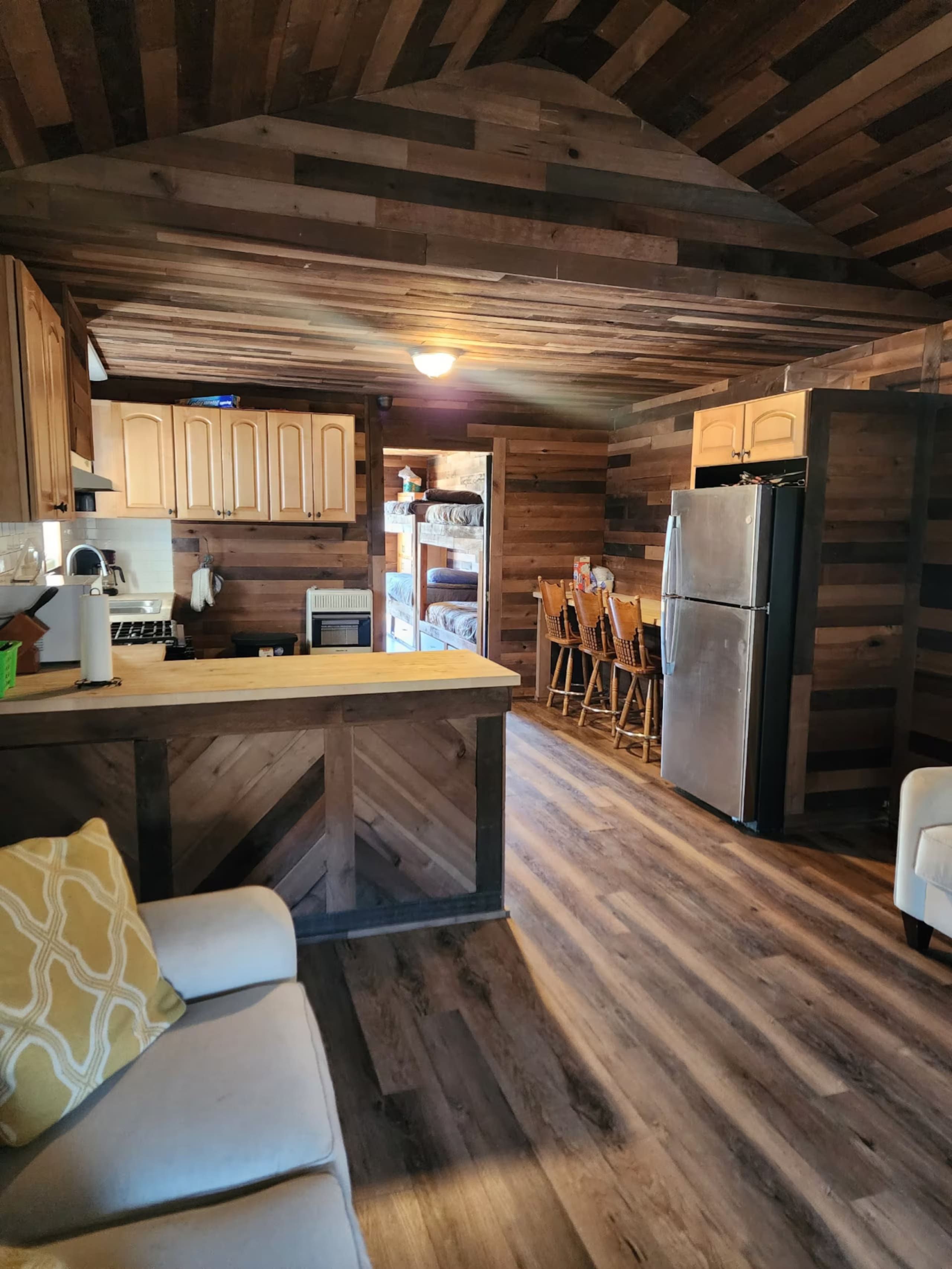 L’intérieur d’un chalet rustique est doté d’un plafond en bois, d’un coin cuisine avec un réfrigérateur, d’une table à manger avec des chaises et d’un canapé agrémenté d’un coussin.