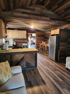 L’intérieur d’un chalet rustique est doté d’un plafond en bois, d’un coin cuisine avec un réfrigérateur, d’une table à manger avec des chaises et d’un canapé agrémenté d’un coussin.