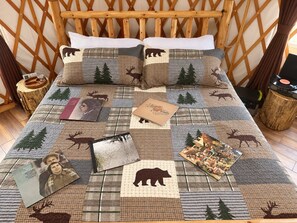 A bed with a patchwork quilt featuring animal prints, surrounded by wooden headboard and nightstands with decorative items. - Lumberjack 20' Yurt, King Bed Glamping in Bend (Bend)