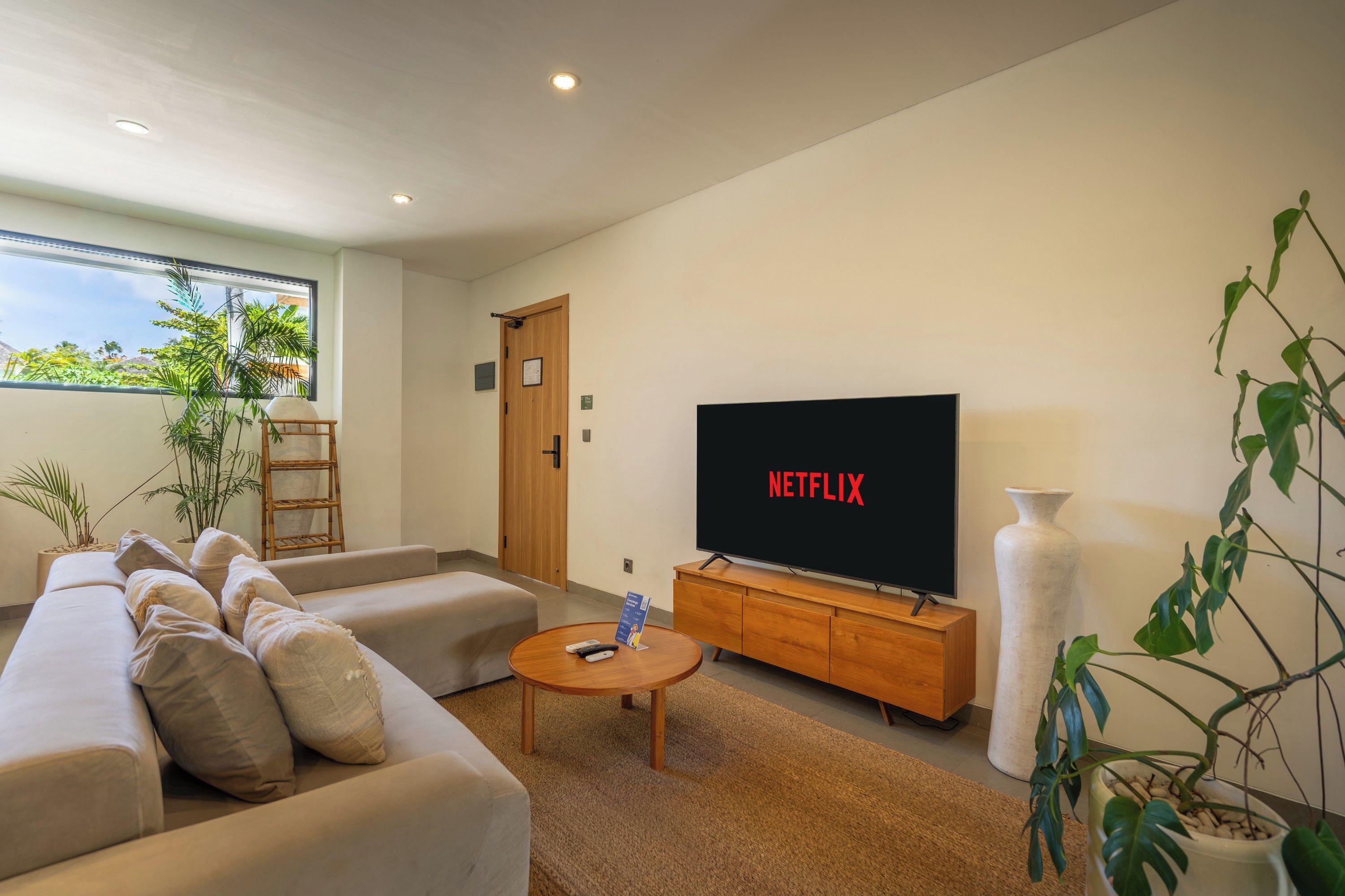 A modern living room with a sofa, a coffee table, a TV displaying 'NETFLIX', and a large window with a view of greenery.