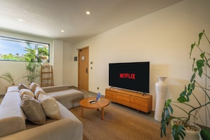 Una sala de estar moderna con un sofá, una mesa de centro, un televisor mostrando 'NETFLIX' y un ventanal con vistas a zonas verdes.