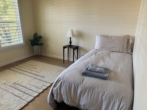 Un dormitorio con una cama, una mesita de noche con lámpara, una ventana con persianas y una planta.