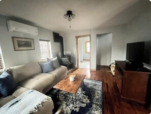 Ein Wohnzimmer mit einer Couch, einem Couchtisch und einem Fernseher. Es gibt einen Holzboden und einen Deckenventilator.