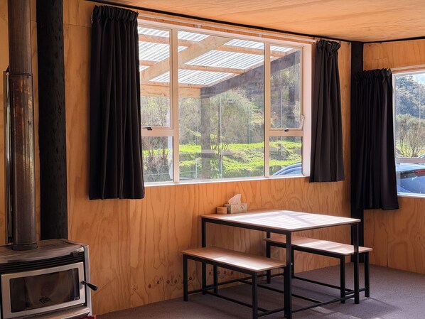 A room with a wooden interior, a table with a bench, a window with curtains, and a wood-burning stove.