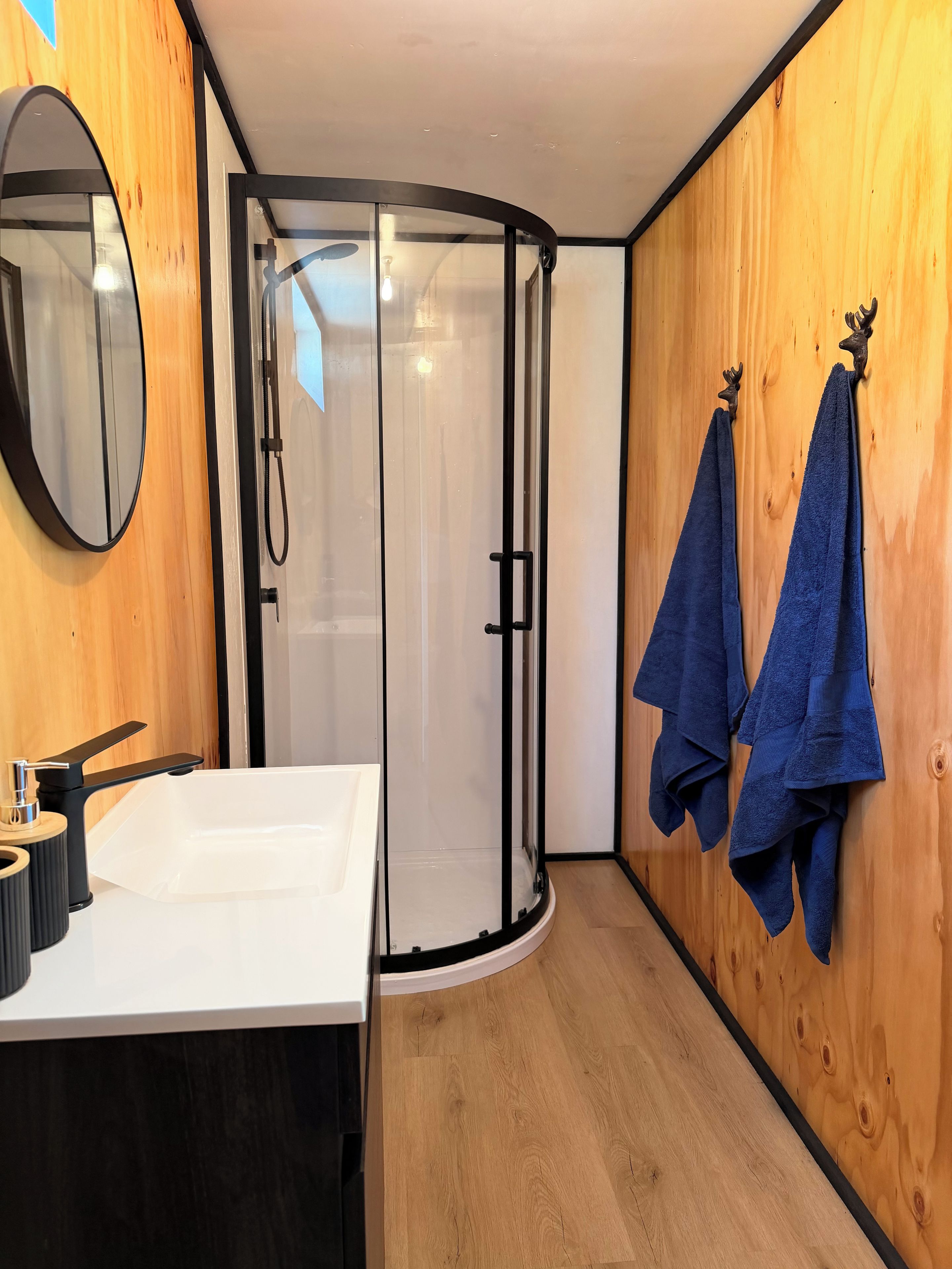 A bathroom with a white sink, a mirror, a shower enclosure, and two towels hanging on a wooden wall.