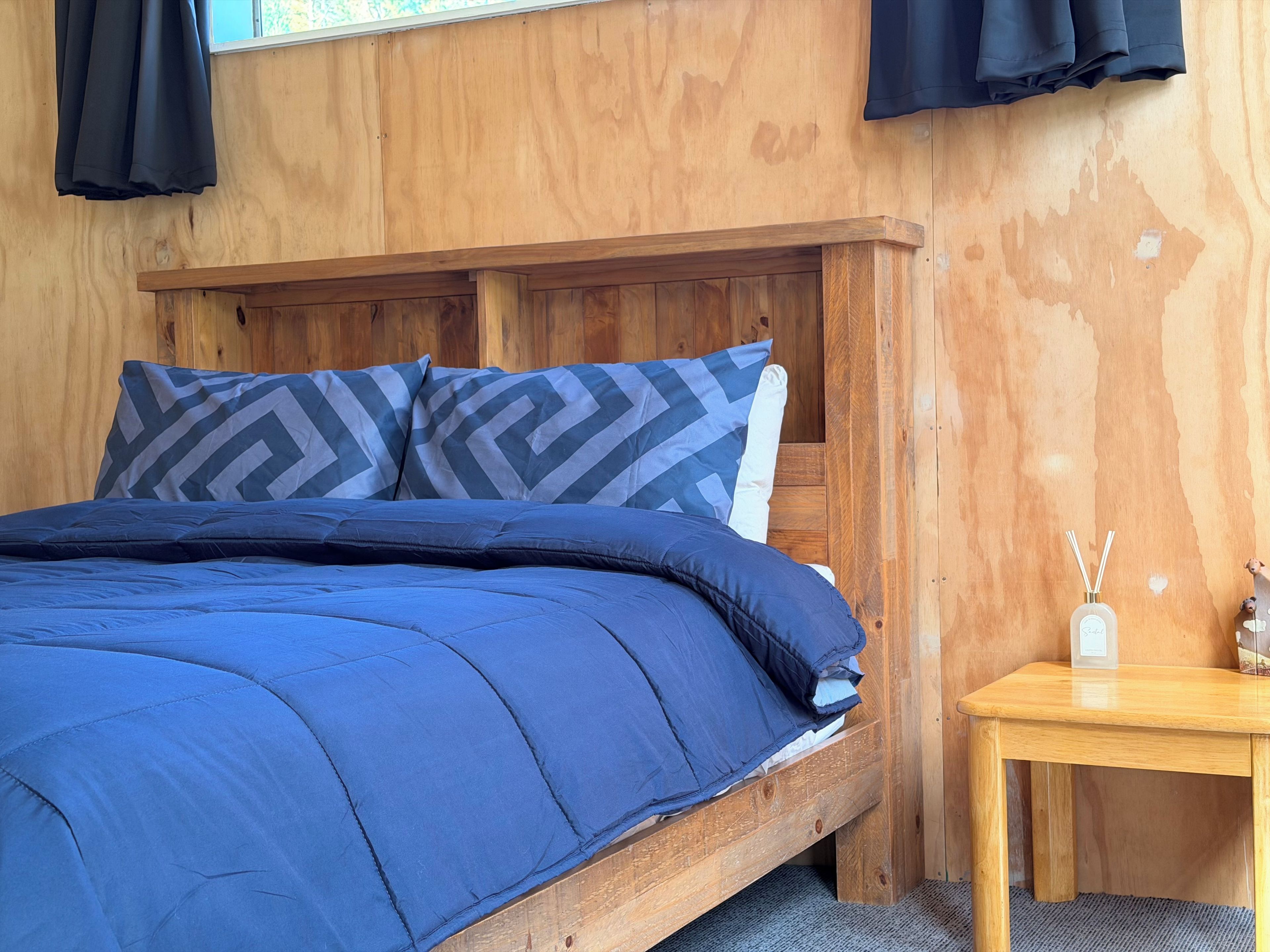 A wooden bed with a blue bedspread and patterned pillows, a small wooden nightstand with a bottle of reed diffuser, and a window with curtains.