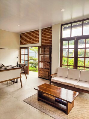A room with a sofa, a coffee table, a dining table, and a view through large windows.
