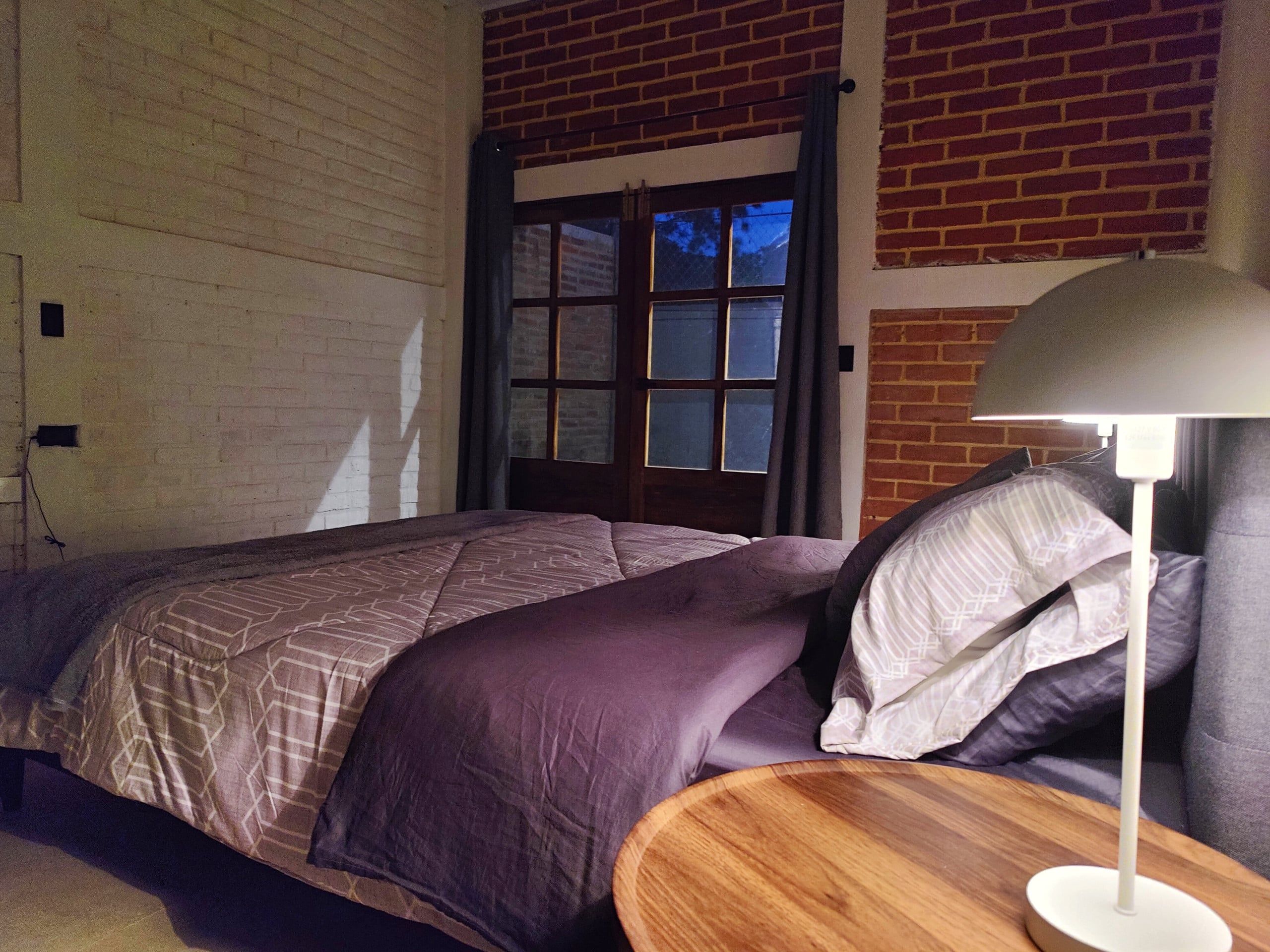 Une chambre à coucher avec un lit, une table de chevet en bois, une lampe et un mur en briques.