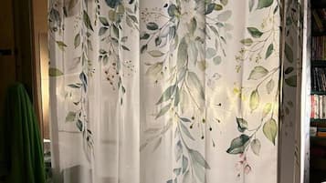 Habitación con cortina blanca estampada con motivos florales, suelo de madera y un ventilador a la derecha.