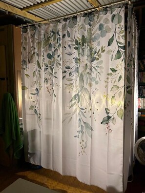Habitación con cortina blanca estampada con motivos florales, suelo de madera y un ventilador a la derecha.