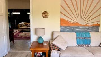 Un salón con sofá, lámpara, una mesita y una pared decorada con un cuadro de sol y montañas.