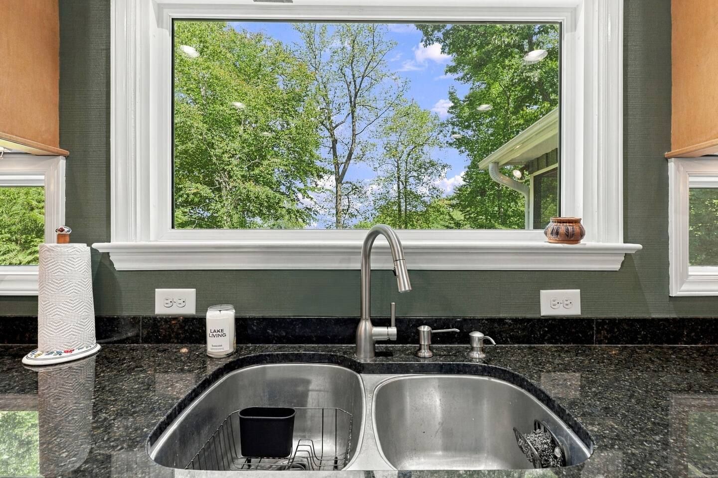 A kitchen with a double sink, a window with a view of trees, and a candle on the counter.