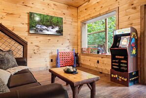 A cozy room with a wooden interior, a couch, a coffee table, a TV mounted on the wall, and a Pac-Man arcade machine. - Penny's Peaceful Pines (Sevierville)