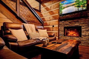 A cozy cabin interior with a leather sofa, a stone fireplace, and a television mounted on the wall. - Penny's Peaceful Pines (Sevierville)