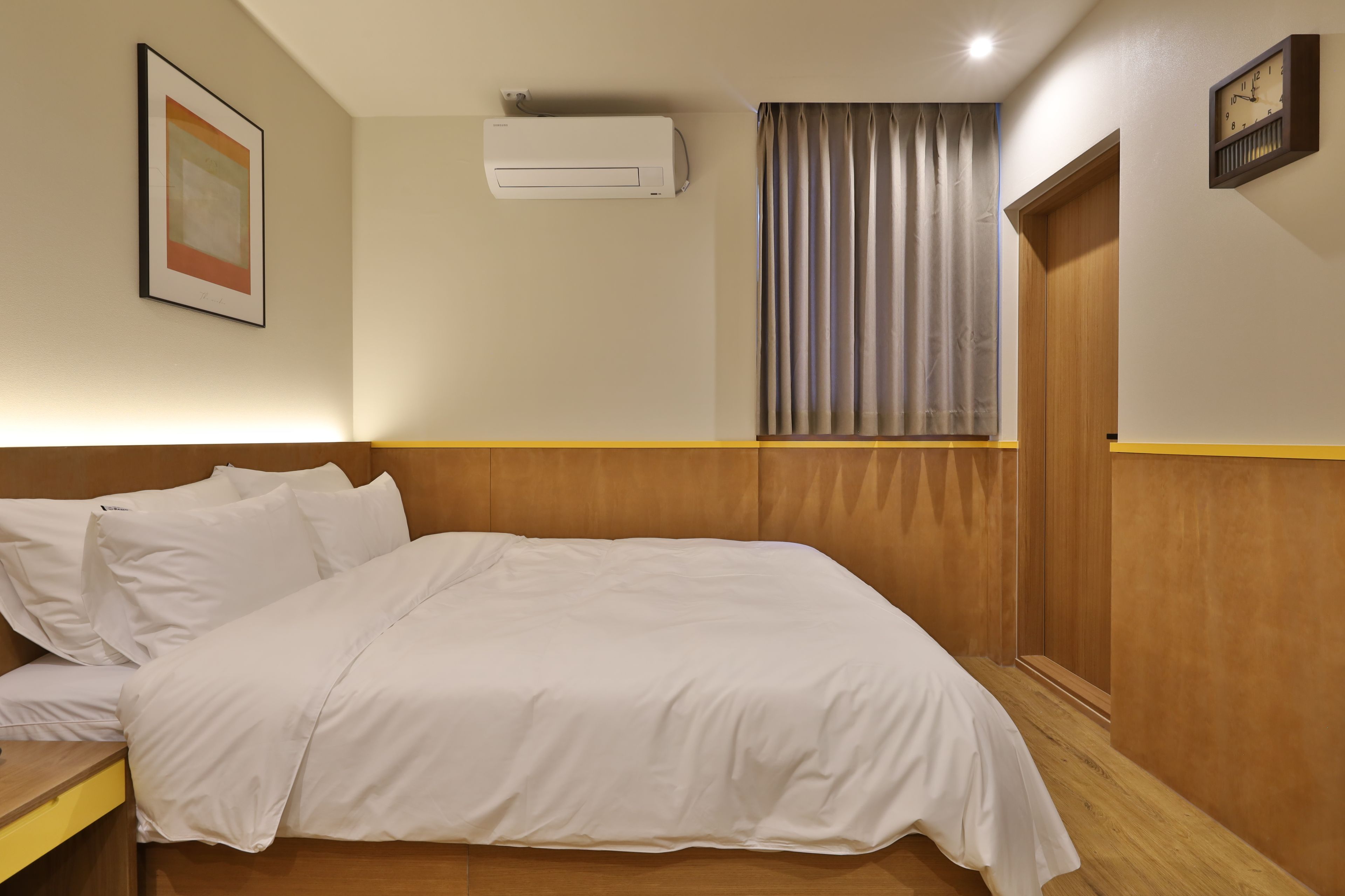 Ein Hotelzimmer mit einem Bett, einem Nachttisch, einer Wandklimaanlage, einem gerahmten Bild und einer Uhr.