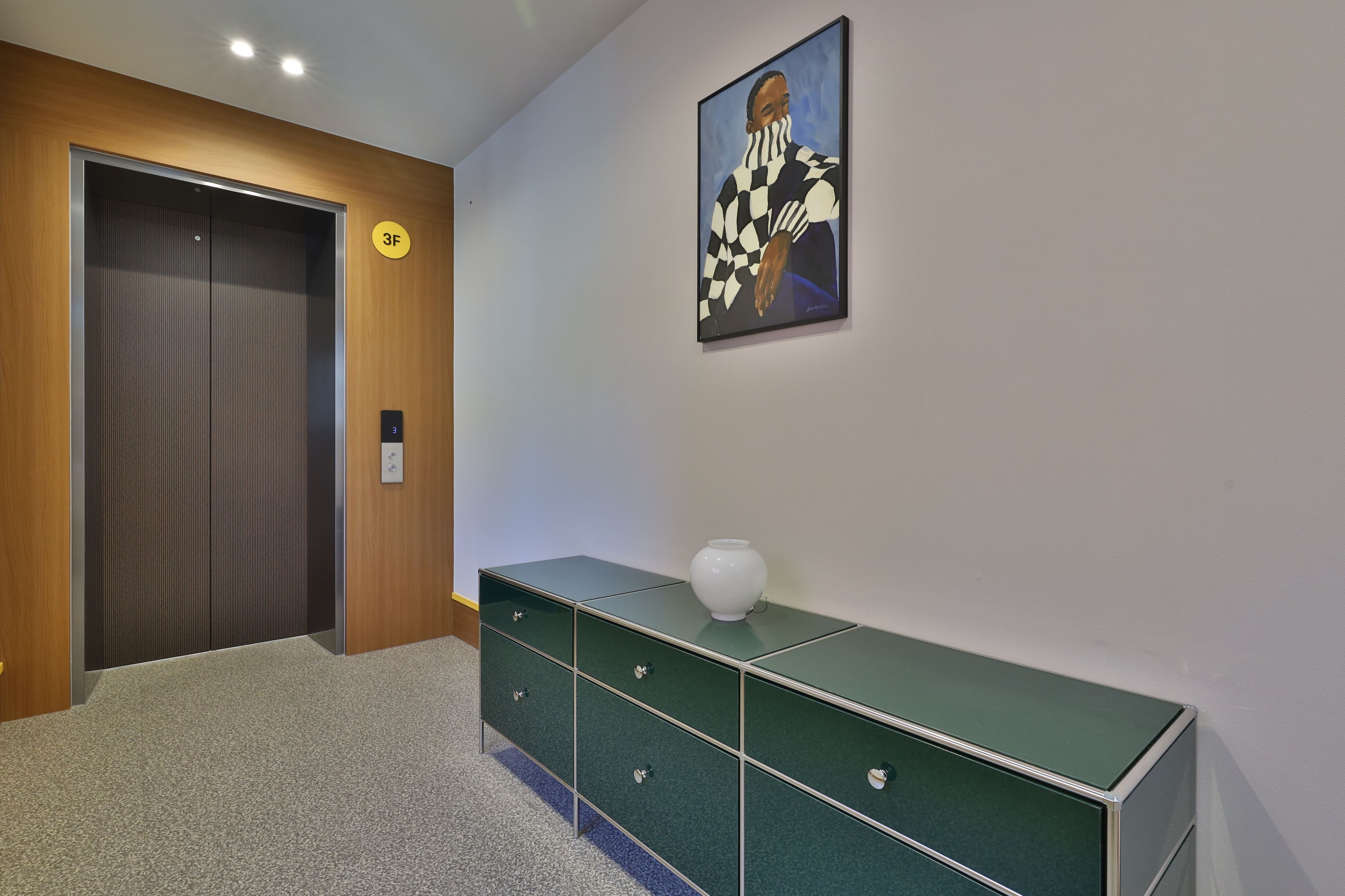 Un pasillo con una puerta de madera, un mueble de almacenamiento verde, un jarrón blanco y un cuadro en la pared.