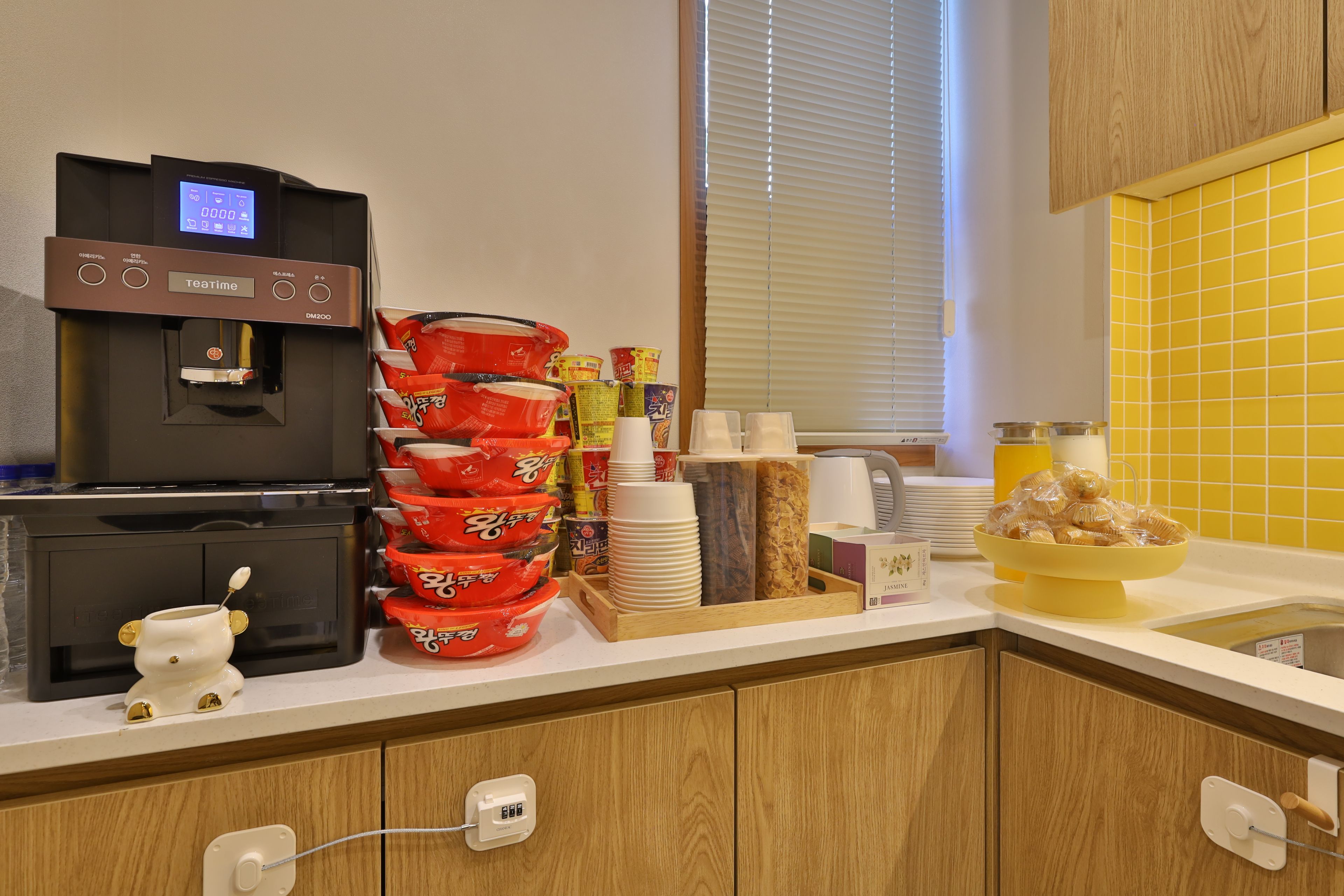 Un coin cuisine équipé d’une machine à café, de piles de gobelets jetables, de condiments et d’un saladier jaune rempli de viennoiseries.