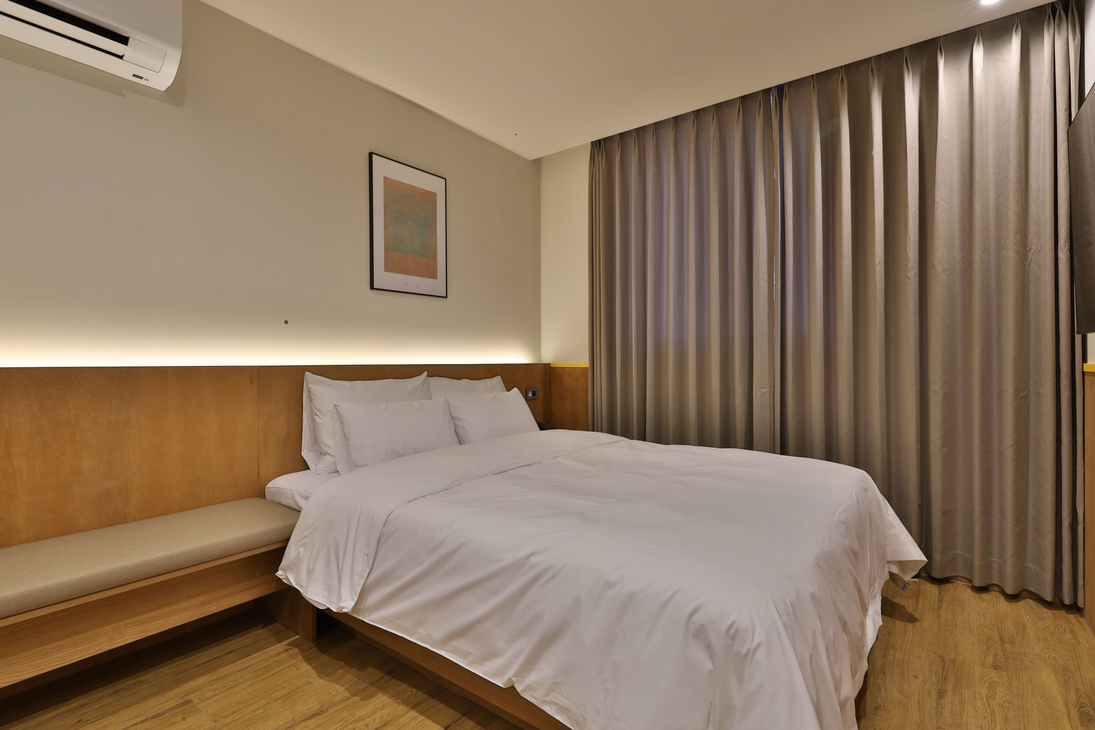 A hotel room with a bed, a bedside table, a wall-mounted air conditioner, a framed picture, and a window with curtains.