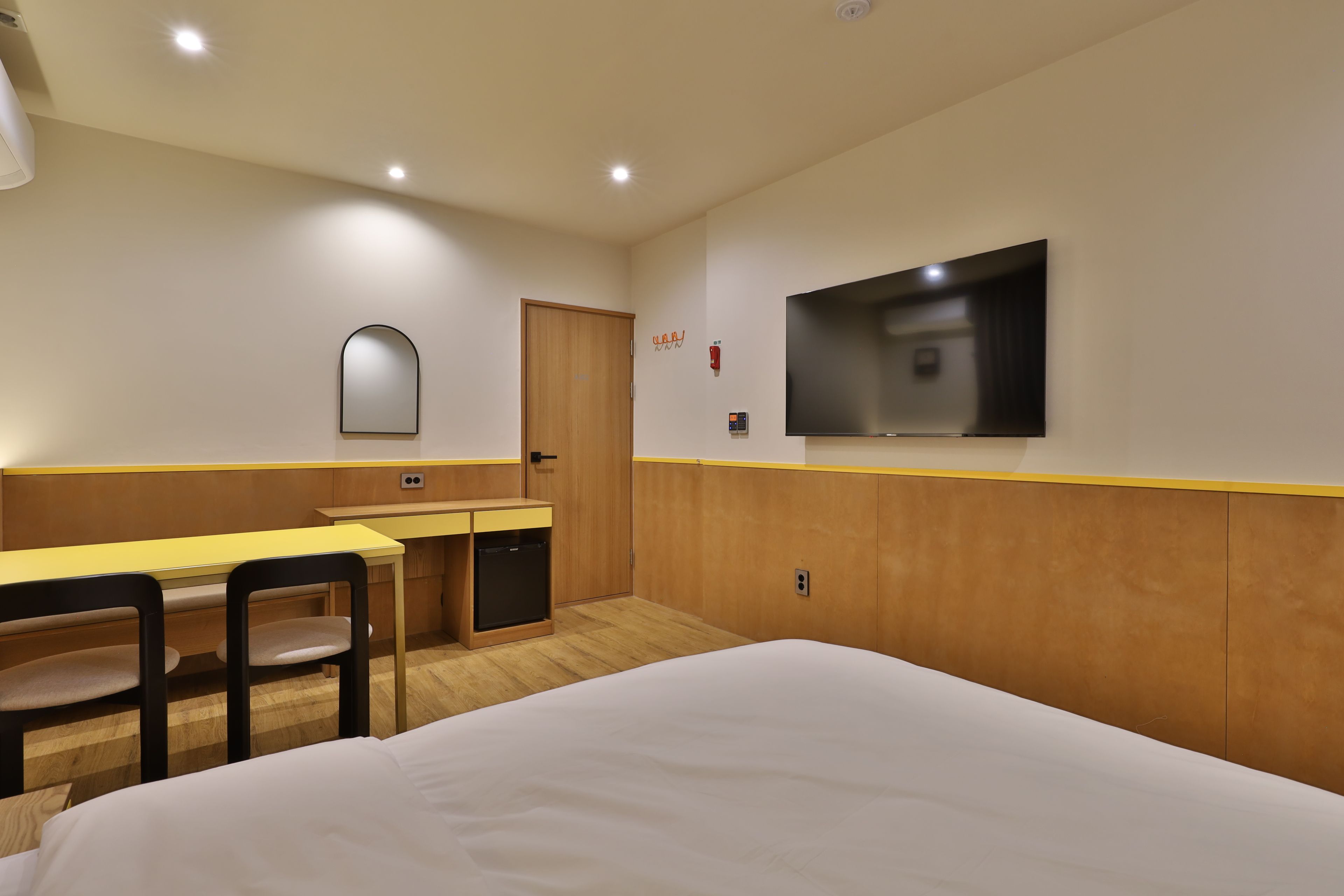 A hotel room with a bed, a desk with a chair, a television mounted on the wall, and a door.