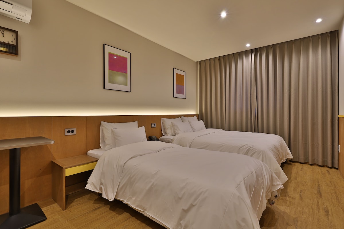 Habitación de hotel con dos camas, una mesita de noche, aire acondicionado instalado en la pared y dos cuadros abstractos enmarcados.