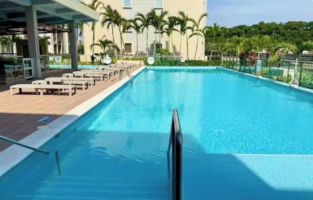 Une piscine dotée de chaises longues et d’un espace abrité, entourée de palmiers et d’un bâtiment.