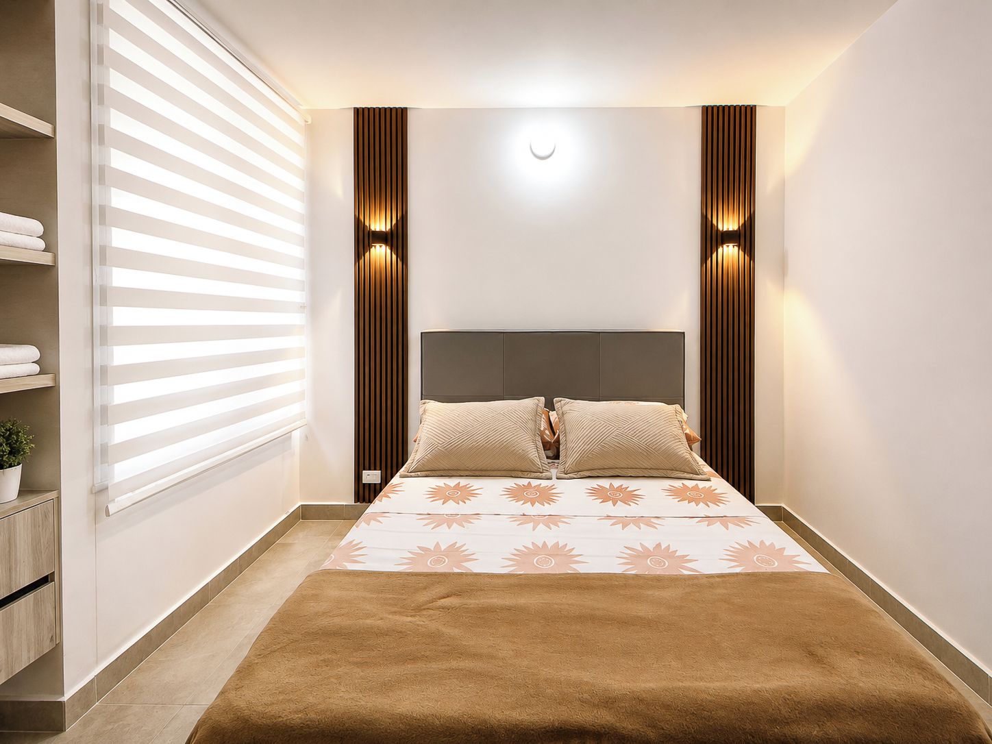 Een slaapkamer met een bed, nachtkastjes, een raam met zonwering en wandlampen.
