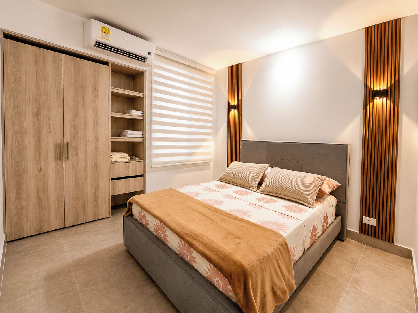 A bedroom with a bed, a wardrobe, a built-in air conditioner, and a window with blinds.