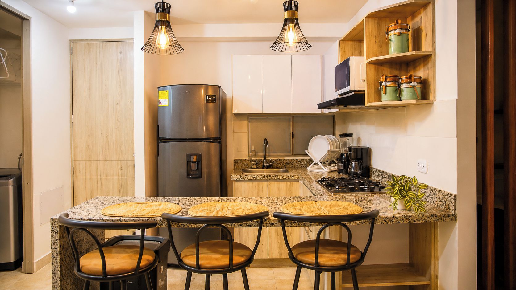 Une cuisine compacte équipée d’un réfrigérateur en acier inoxydable, d’un four à micro-ondes et d’une cuisinière. On y trouve trois tabourets de bar avec des assises en osier et un comptoir en granit.