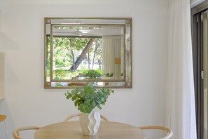 A dining area with a round table, a white vase with green plants, and a mirror reflecting a view of trees and a building. - Kananda Riverside, Noosa Heads (Noosa Heads)