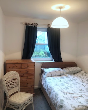 A bedroom with a bed, a wooden dresser, a wicker chair, a window with curtains, and a ceiling light. - Cosy flat 1 min from Union Street, perfect for 2 or a small family (Aberdeen)