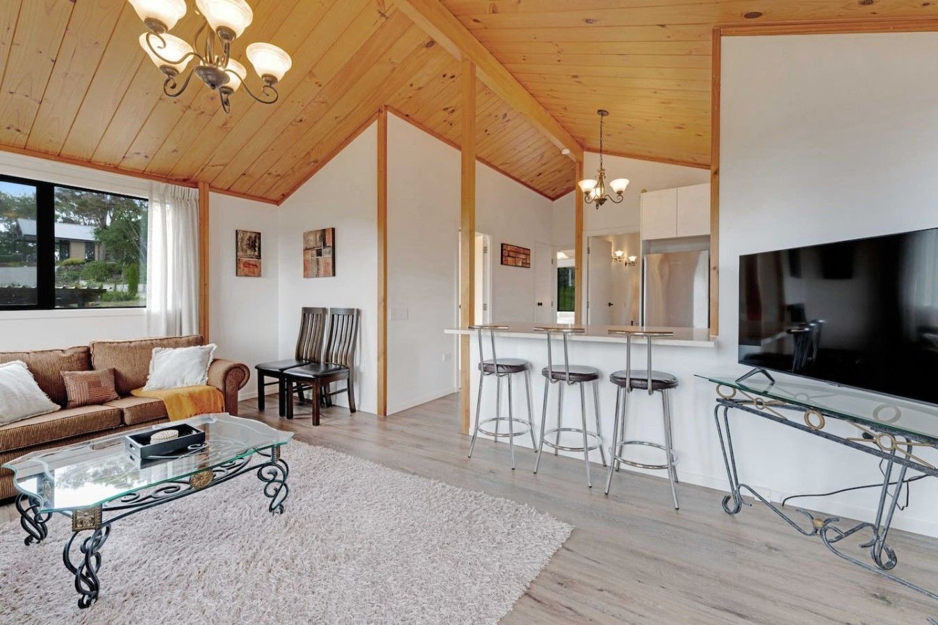 A modern living room with a sofa, coffee table, and a TV. There are three bar stools and a glass-top table. The room has wooden ceilings and a large window.