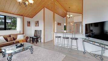 A modern living room with a sofa, coffee table, and a TV. There are three bar stools and a glass-top table. The room has wooden ceilings and a large window.