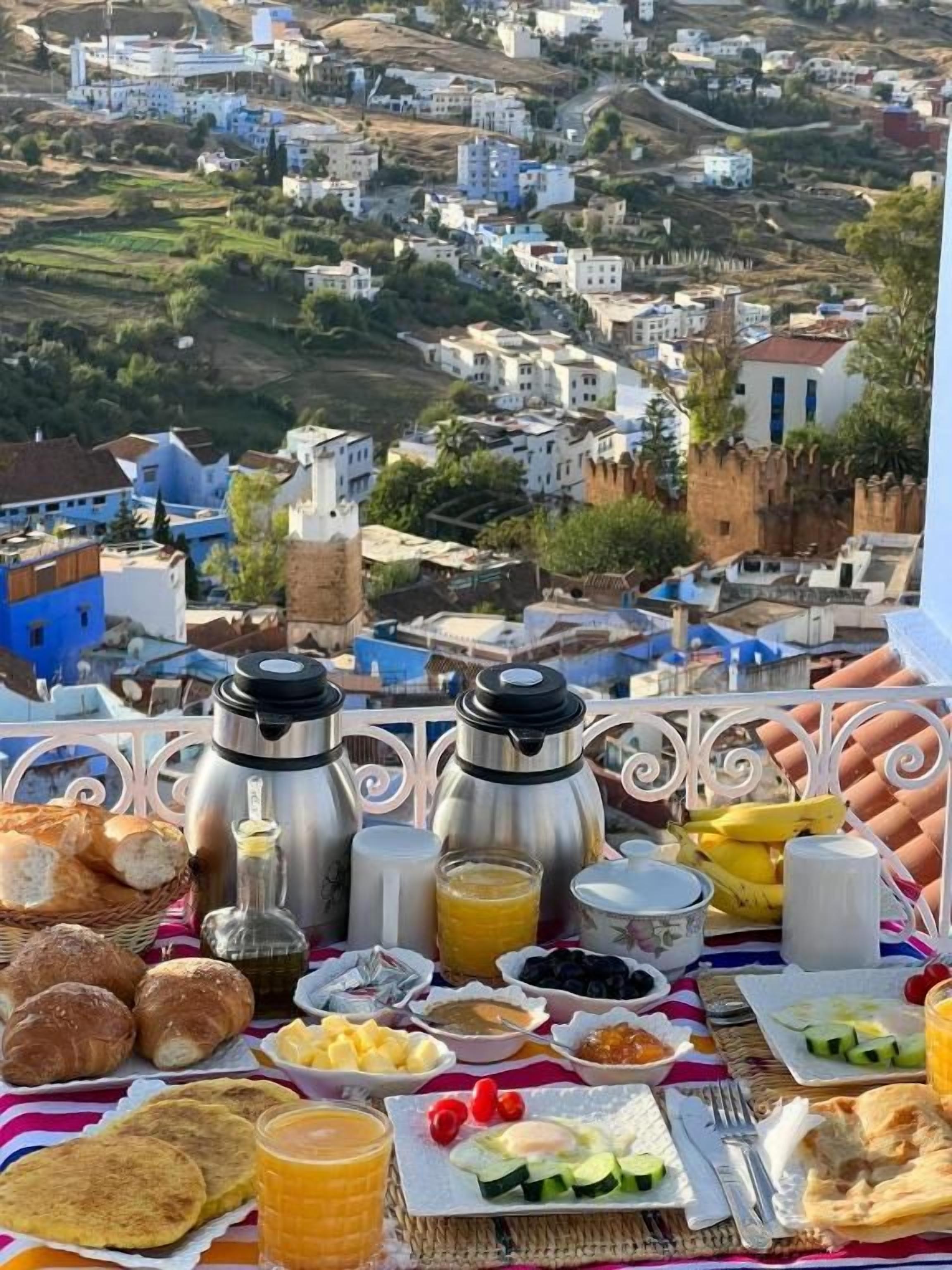 A breakfast setup with various food items and two coffee makers on a table, overlooking a town with buildings and greenery.