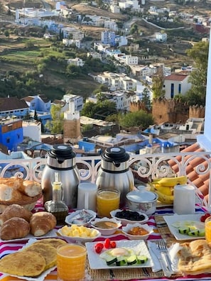 Preparación de desayuno con diferentes alimentos y dos cafeteras sobre una mesa, con vistas a un pueblo con edificios y vegetación.