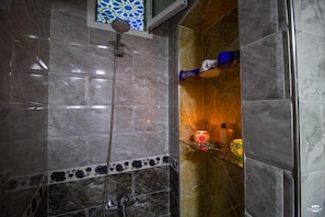 Un baño con ducha, azulejos de mármol y un estante de vidrio con artículos de aseo.