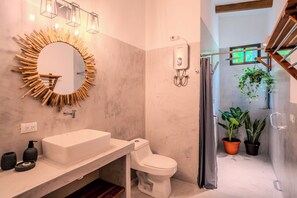Ein Badezimmer im modernen Stil, mit einem weißen Waschbecken, einer Toilette und einem Spiegel mit einem markanten Holzrahmen. Sichtbar sind außerdem Blumentöpfe und ein Duschbereich.