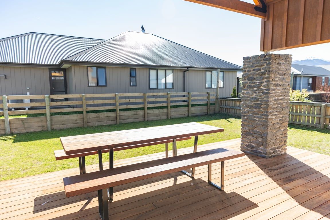 A wooden deck with a picnic table, a stone pillar, and a fenced area with a building in the background.