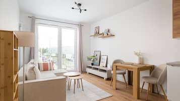 A modern living room with a sofa, a small round table, and a dining table with chairs. There is a ceiling light fixture and a view of the outside through large windows.