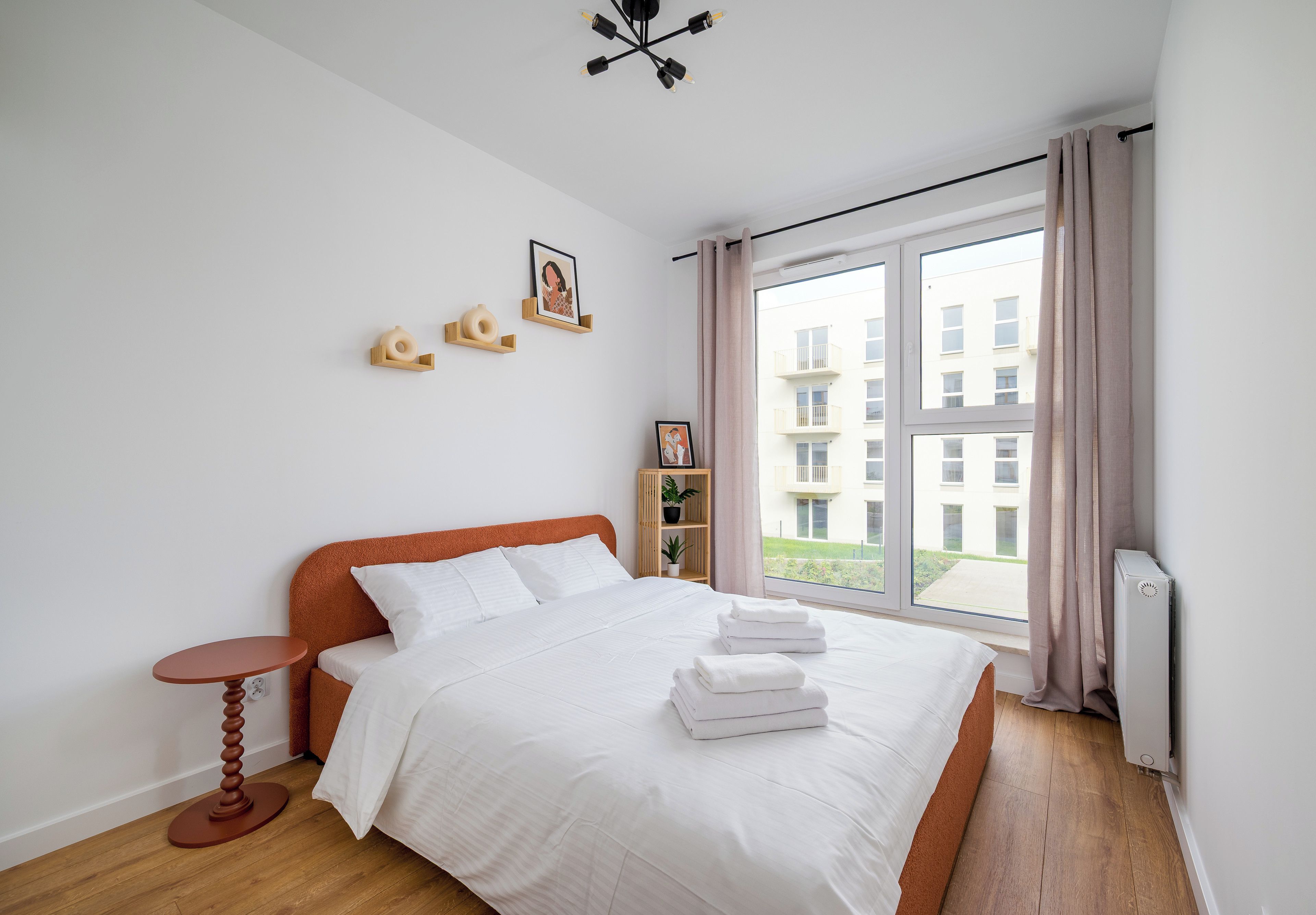 Un dormitorio con una cama, una mesita de noche, una ventana con cortinas y vistas a edificios.