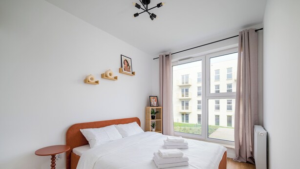 Un dormitorio con una cama, una mesita de noche, una ventana con cortinas y vistas a edificios.