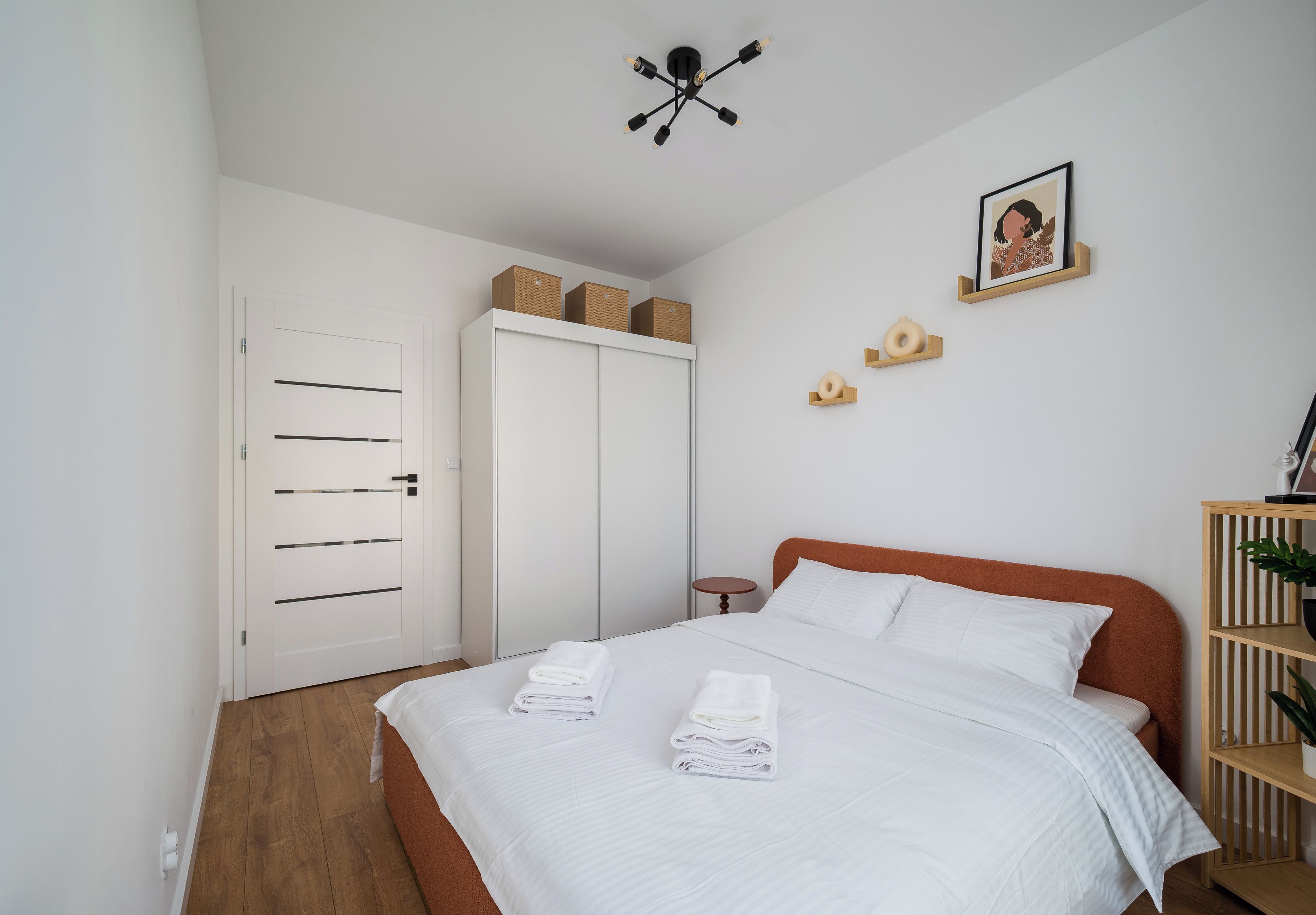 A bedroom with a bed, white walls, a wardrobe, a ceiling light fixture, a framed picture, and a small wooden shelf.